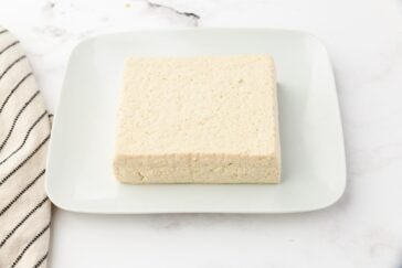 A block of tofu sits on a white square plate with a striped cloth napkin beside it. The background is a smooth white surface.