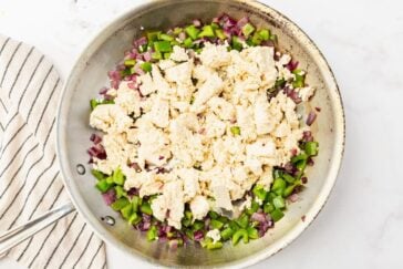 A skillet containing crumbled tofu, green bell peppers, and red onions. The ingredients are uncooked and spread evenly in the pan. A striped kitchen towel is partially visible on the side.
