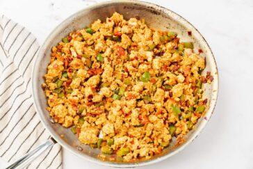 A skillet filled with tofu scramble mixed with diced green peppers, onions, and tomatoes, on a light surface. A striped cloth is partially visible beside the skillet.