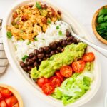 A dish filled with white rice, black beans, seasoned tofu, guacamole, halved cherry tomatoes, and shredded lettuce. A spoon is placed on the side. Bowl of fresh cilantro nearby on a white surface.