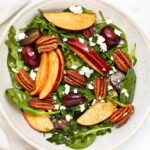 A salad in a bowl with spinach, sliced apples, pecans, grapes, and crumbled feta cheese, all tossed in a dressing.