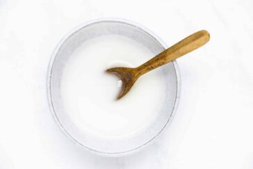 A bowl of white liquid with a rustic brown wooden spoon resting inside, set against a plain white background.