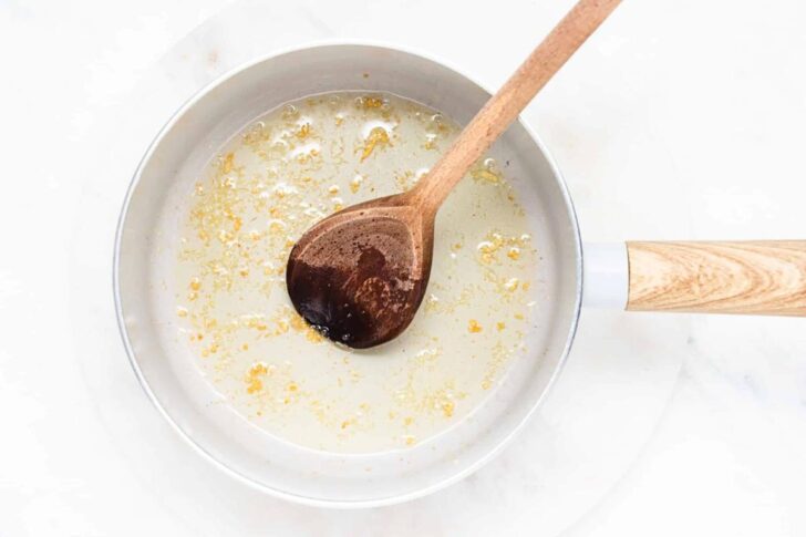A saucepan with melted butter and minced garlic is being stirred by a wooden spoon, sitting on a white surface.