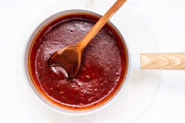 A saucepan filled with rich, red barbecue sauce. A wooden spoon is resting inside, indicating the sauce has been freshly stirred. The sauce has a smooth texture and is placed on a white background.