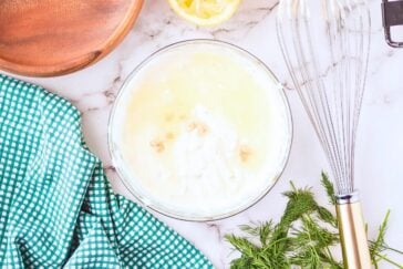 A bowl of creamy white dip sits on a marble countertop. Nearby are a whisk, a wooden plate, a checkered green cloth, dill sprigs, and a slice of lemon, suggesting a fresh and homemade cooking setup.