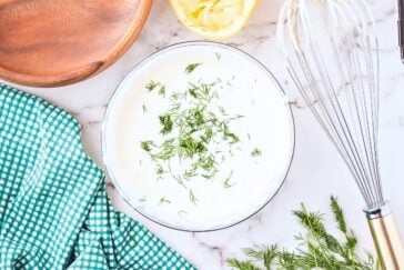 A bowl of creamy sauce garnished with dill is surrounded by a whisk, a cut lemon, a wooden bowl, and a green checkered cloth on a marble countertop. Fresh dill is scattered nearby.