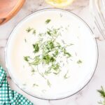 A bowl of creamy white sauce garnished with fresh dill on a marble surface. A whisk, a wooden bowl, and a halved lemon are partially visible around the bowl. A green checkered cloth is on the side.