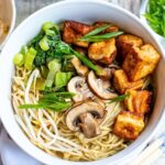 A bowl of noodle soup topped with fried tofu cubes, mushrooms, bean sprouts, bok choy, and sliced green onions. The noodles are submerged in a light broth, creating a colorful and appetizing dish.