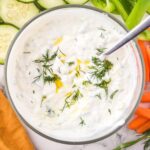 A bowl of creamy yogurt dip garnished with fresh dill and olive oil. Surrounding the bowl are fresh vegetables, including sliced cucumbers, carrots, and green bell peppers, partly visible. A spoon rests in the dip.