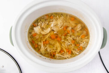A white slow cooker filled with chicken soup, featuring shredded chicken, chopped carrots, celery, and onions in a clear broth. The lid is placed beside the cooker on a white surface.