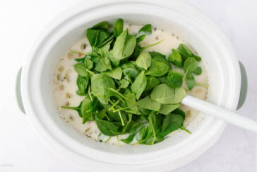 A white crockpot filled with creamy soup base, topped with fresh spinach leaves. A white spoon is resting inside, ready to stir. The spinach appears vibrant against the pale liquid, indicating the dish is in preparation.
