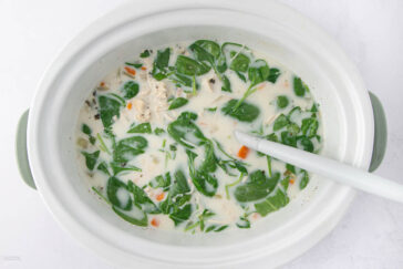 A white pot filled with creamy chicken soup, featuring pieces of chicken, diced carrots, and fresh spinach leaves. A ladle rests inside, and the soup has a mixture of creamy white broth and green herbs.