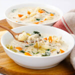 Two bowls of creamy chicken gnocchi soup with carrots, celery, and spinach, served on a wooden board. A spoon is lifting a portion from one bowl. A striped napkin is nearby, adding a cozy touch.