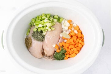 Raw chicken breasts in a slow cooker with chopped celery, carrots, onions, bay leaf, and sprinkled herbs like thyme and parsley, ready for cooking.