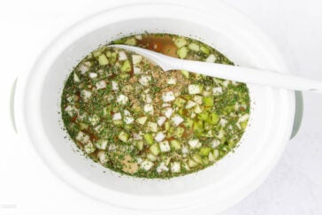 A white slow cooker filled with a seasoned broth mixture. The broth contains chopped green vegetables and herbs. A white ladle rests inside the cooker, partially submerged in the liquid.
