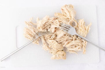 Shredded cooked chicken on a white cutting board with two forks resting on top.