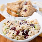 A bowl of chicken salad with grapes, celery, and nuts on a wooden surface. In the background, two whole grain sandwiches filled with the same chicken salad are placed on a white plate with a striped napkin.