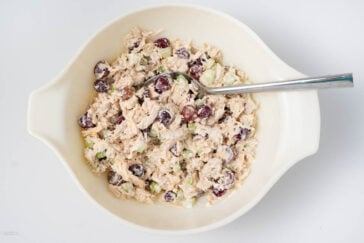 A bowl of chicken salad with grapes, celery, and a creamy dressing, mixed and ready to serve. A metal spoon is placed in the bowl for serving. The dish is set against a plain white background.