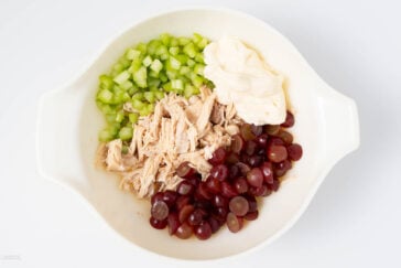 A bowl containing chopped celery, shredded chicken, sliced red grapes, and a dollop of mayonnaise. The ingredients are arranged separately and ready to be mixed for a salad.