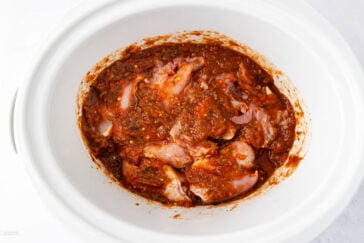 Raw chicken pieces marinated in a thick, red-brown sauce inside a white slow cooker, ready for cooking.