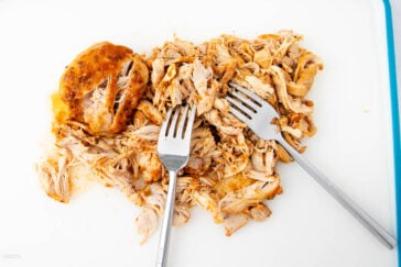 Shredded cooked chicken on a cutting board with two metal forks. The chicken is seasoned and appears juicy.