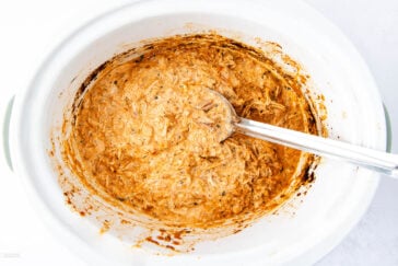 A slow cooker filled with creamy shredded chicken, mixed with a reddish sauce. A metal spoon rests inside, ready to serve. The mixture looks rich and flavorful, with flecks of seasoning visible throughout.