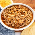 A white oval dish filled with shredded Mexican chicken, seasoned with spices, sits on a wooden surface. Nearby are corn tortillas, a small bowl of shredded cheese, and a dark patterned cloth.