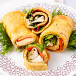 Four rolled omelette wraps filled with slices of chicken, lettuce, and tomato are placed on a decorative white plate with red circular patterns.