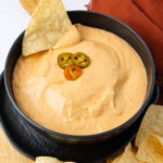 A black bowl filled with creamy nacho cheese dip, topped with sliced jalapeños. A tortilla chip is dipped into the cheese, and more chips are scattered around. The bowl is placed on a wooden surface with an orange napkin nearby.