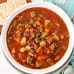 A bowl of hearty vegetable and beef soup with chunks of ground beef, carrots, celery, potatoes, green beans, tomatoes, and white beans. A few saltine crackers are on the side. The soup is in a white bowl on a checkered cloth.