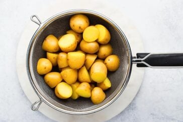 A metal colander filled with halved small yellow potatoes sits on a round white marble surface. The potatoes have a smooth, golden-brown skin and are evenly cut. The background is a light, textured surface.