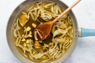 A pot filled with sliced onions, ginger pieces, and bay leaves simmering in broth. A wooden spoon rests in the pot, stirring the ingredients. The pot has a light blue handle and sits on a light surface.