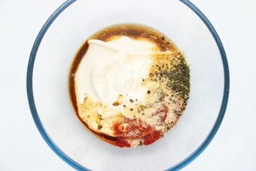 A clear bowl contains a mixture of mayonnaise, soy sauce, spices, and herbs on a white background. The ingredients are partially blended, showcasing creamy and liquid textures with visible specks of seasoning.