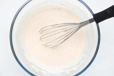 A whisk resting in a clear glass bowl containing creamy, beige batter. The mixture has small visible specks and is smooth in texture, suggesting it might be a sauce or dressing in preparation. The bowl is on a light-colored surface.