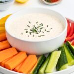 A white bowl filled with creamy ranch dip topped with chopped chives sits on a platter. Surrounding the bowl are colorful vegetable sticks, including orange carrots, green cucumbers, and red and yellow bell peppers.
