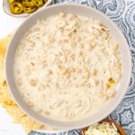 A bowl of creamy white chicken chili garnished with shredded chicken and white beans, surrounded by tortilla chips, a small dish of jalapeños, and shredded cheese on a patterned tablecloth.