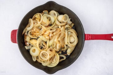 A red-handled skillet filled with sautéed onion slices, lightly browned, sitting on a white surface.