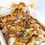 A white casserole dish filled with French onion chicken. The dish features melted cheese and caramelized onions, with herbs sprinkled on top. A spoon lifts a portion, showcasing the cheese pull.