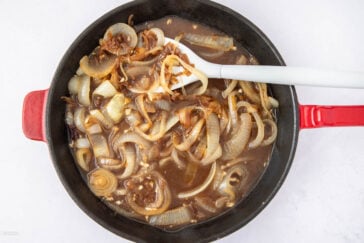 A skillet filled with caramelized onions and sauce, being stirred with a white spatula. The onions are sliced and submerged in a brown liquid. The skillet has a red handle and rests on a white background.