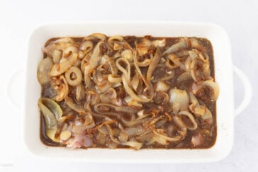 A rectangular white dish filled with sliced onions in a dark liquid, possibly sauce or marinade. The dish shows a variety of onion textures and shades, from translucent to lightly browned.