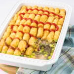 A casserole dish filled with tater tots arranged neatly on top of a mixture of vegetables and beef in a creamy sauce. The tater tots are topped with zigzagging lines of ketchup. A plaid cloth is partly visible beside the dish.