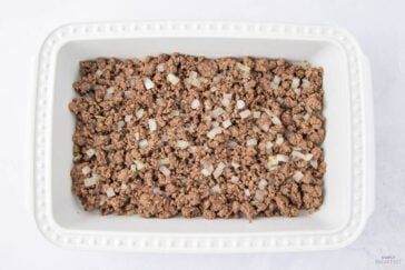 A rectangular white ceramic dish containing cooked ground beef mixed with diced onions, placed on a light surface. The beef is evenly spread across the dish, showing a textured and savory appearance.