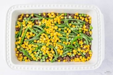 A white baking dish filled with layers of mixed vegetables on top of ground beef. The veggies include green beans, corn, and peas, creating a colorful mix. The edges of the dish are adorned with a dotted design.