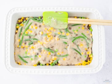 A rectangular white baking dish filled with a layered casserole. It contains ground meat, corn, peas, and green beans, topped with a creamy sauce. A green spatula rests on top of the casserole, partially covered in sauce.