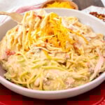 A white bowl filled with creamy chicken spaghetti topped with shredded cheddar cheese. The dish is garnished with diced tomatoes and features a fork lifting a portion of the pasta. A small bowl of cheddar and red pepper flakes are in the background.