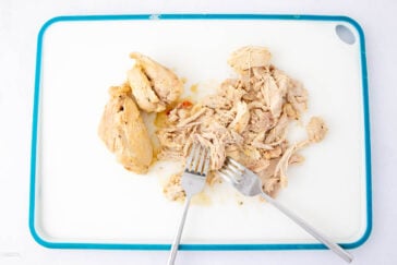 Shredded cooked chicken and two forks on a white cutting board with a blue border.