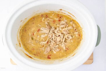 A white slow cooker filled with creamy chicken and vegetable stew. Shredded chicken is visible on top, surrounded by a thick, golden broth with bits of red and green vegetables. The cooker sits on a wooden surface.