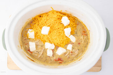 A slow cooker filled with a creamy chicken soup mixture. Shredded cheddar cheese and cubes of cream cheese are placed on top. The soup contains visible pieces of chicken and diced tomatoes, with herbs mixed in.