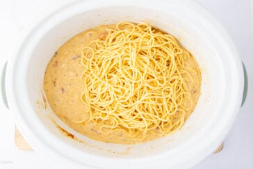A crockpot filled with creamy spaghetti topped with shredded cheese. The pasta is partially mixed with a cheese and tomato sauce, creating a rich, inviting texture. The crockpot sits on a light-colored surface.