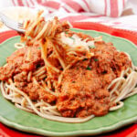 A plate of spaghetti topped with rich meat sauce on a green plate. A fork is lifting a portion of the pasta. The dish is set against a red placemat and a striped cloth, adding a colorful background to the meal.